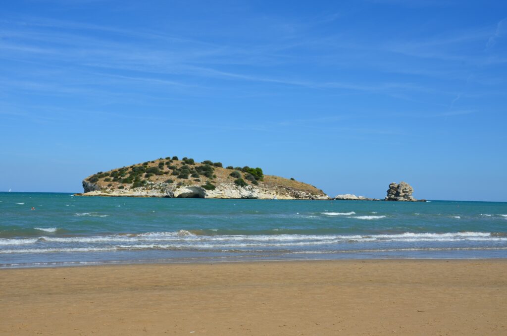 Yellow sandy beach by the sea, in which you can see a small island covered with trees, next to it is a large rock.