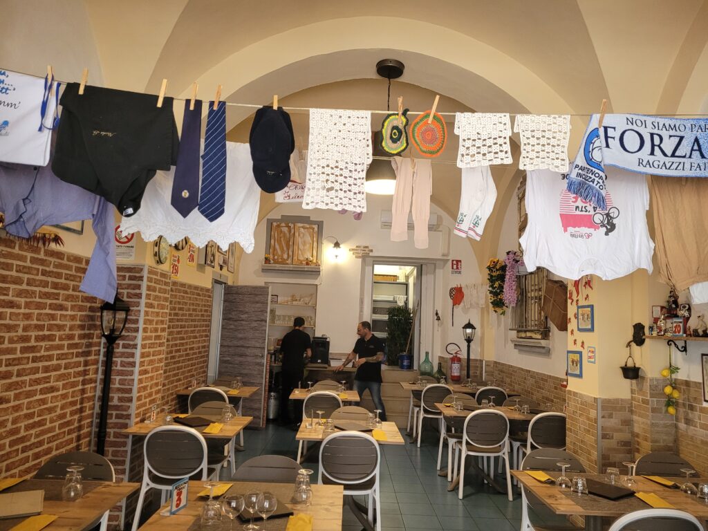A restaurant with wooden tables and brown chairs on which people sit, clothes hanging on lines above their heads and between the walls.