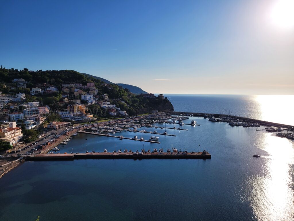 On the shore of the coast, on green hills, there is a sea with small houses, in the very blue sea there are piers and small boats.