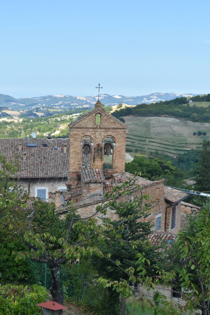 A stone belfry with two bells, a metal cross, and a tiled roof stands among the greenery.