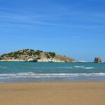 Yellow sandy beach by the sea, in which you can see a small island covered with trees, next to it is a large rock.