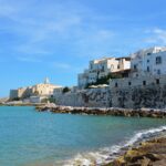 The rocky shore of the blue sea, above it is a wall behind which stand white buildings.