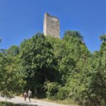 a bright road at a bend, along which two people are walking, behind them there are trees and above them you can see the ruins of a castle tower