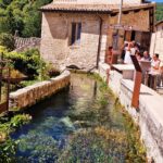 A stream of water flows through a stone canal, at the end of which stands an old stone house. People stand on one side of the canal, and trees on the other.
