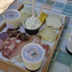 On a wooden tray with handles there are pieces of cheese, slices of cold meats and yogurt in cups.