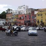 A scooter and a white car drive down the street alongside red and yellow houses.