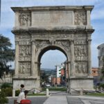 A tall ancient triumphal arch stands in the street, and in front of it a man sits on a bench.
