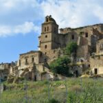 Behind a metal chain-link fence on a green hill stand the ruins of stone houses and a stone church.