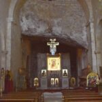 Two rows of wooden benches stand in front of the altar, a large Orthodox cross hangs from the ceiling