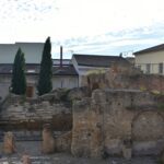 The ruins of an ancient building stand against the background of residential buildings and two tall trees.