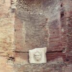 A high wall made of orange brick on which you can see a white mask carved in white marble.