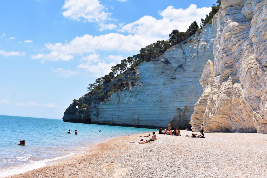 on a pebble Vignanotica Beach under a white cliff covered with trees, several people are sitting on a blanket, three more people are swimming in the blue sea, only their heads are visible