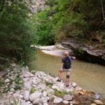 On the stone bank of the river stands a woman in shorts, with a backpack on her back, taking photos.