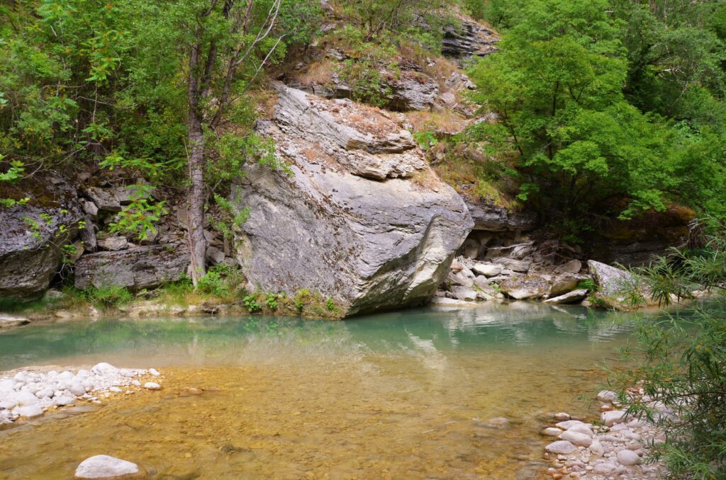Trekking in Abruzzo and a huge boulder lies in a yellow-blue mountain river, with trees and plants growing on its banks