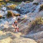 A woman with blond hair, wearing shorts and pink sandals is walking up a beige, dry, cracked hill.