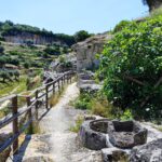The prehistoric stone ruins of the structure lie along a beaten dirt road, with a wooden railing running along one side.