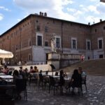 In the square there are people sitting at café tables, and behind them stands a large stone building.