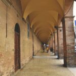 Yellow old stone cloisters supported by columns above the passageway where people are walking.