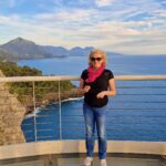 a woman in a black blouse, pink headband and jeans stands on a glass terrace by a metal railing. Behind her you can see the seashore and white clouds in the sky.
