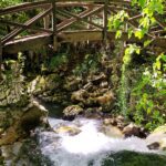 Greenery covers the banks of a foaming river, over which a wooden bridge rises.