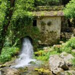 An old stone building from which a stream of water flows and falls over the stones into a river flowing among the greenery.