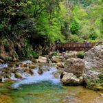 Blue water in the river flows among the rocky shore, in the background you can see a wooden bridge.