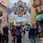 People stand on the street overlooking the sea, among the old houses, colorful circles hanging above their heads.