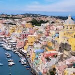 yellow, blue, white and orange houses stand in a port by the sea, where you can see boats. Behind the houses you can see the sea