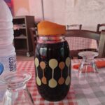 On a table covered with red-and-white checked oilcloth sits a glass jug with an orange lid and yellow and orange patterns. Next to it is a bottle of water, and two wine glasses sit upside down on paper napkins. Behind the table, you can see the back of a chair, and on the wall, furniture.
