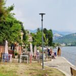 Along the lake's shore runs an alley, fenced on one side with a metal railing, overlooking the lake. In the background are green mountains and boats on the lake. Several people sit on a bench by the alley, and a woman in a sari stands by the railing. Behind the people on the bench are colorful restaurant tables, over which twinkling lights hang.