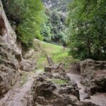 Green trees grow on a small hill. The hill is covered with grass, and wooden steps lead down the hill, ending at large boulders lying beside a well-trodden path. On one side of the path is a small rock bridge with trees growing on it.